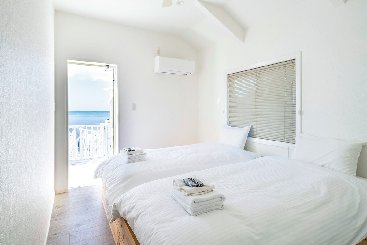A bedroom with two beds, white bedding, a view of the sea through a balcony door, and an air conditioning unit on the wall.