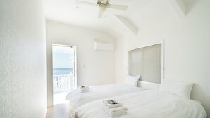 A bedroom with two beds, a ceiling fan, an air conditioning unit, a window with blinds, and a view of the sea. - Sunset Resort Kanutan (Motobu)