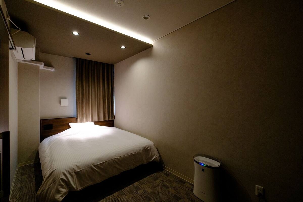 A hotel room with a bed, a waste bin, a ceiling light, and a wall-mounted air conditioning unit.
