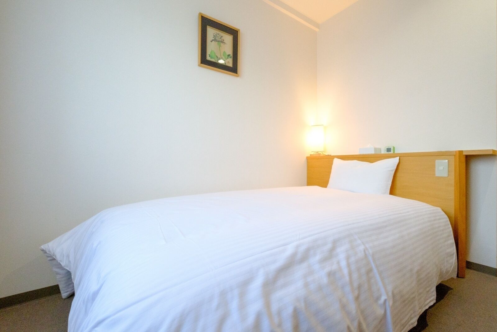 Een enkel bed met een houten hoofdbord, een nachtkastje met een lamp en een lijst met een afbeelding aan de muur.