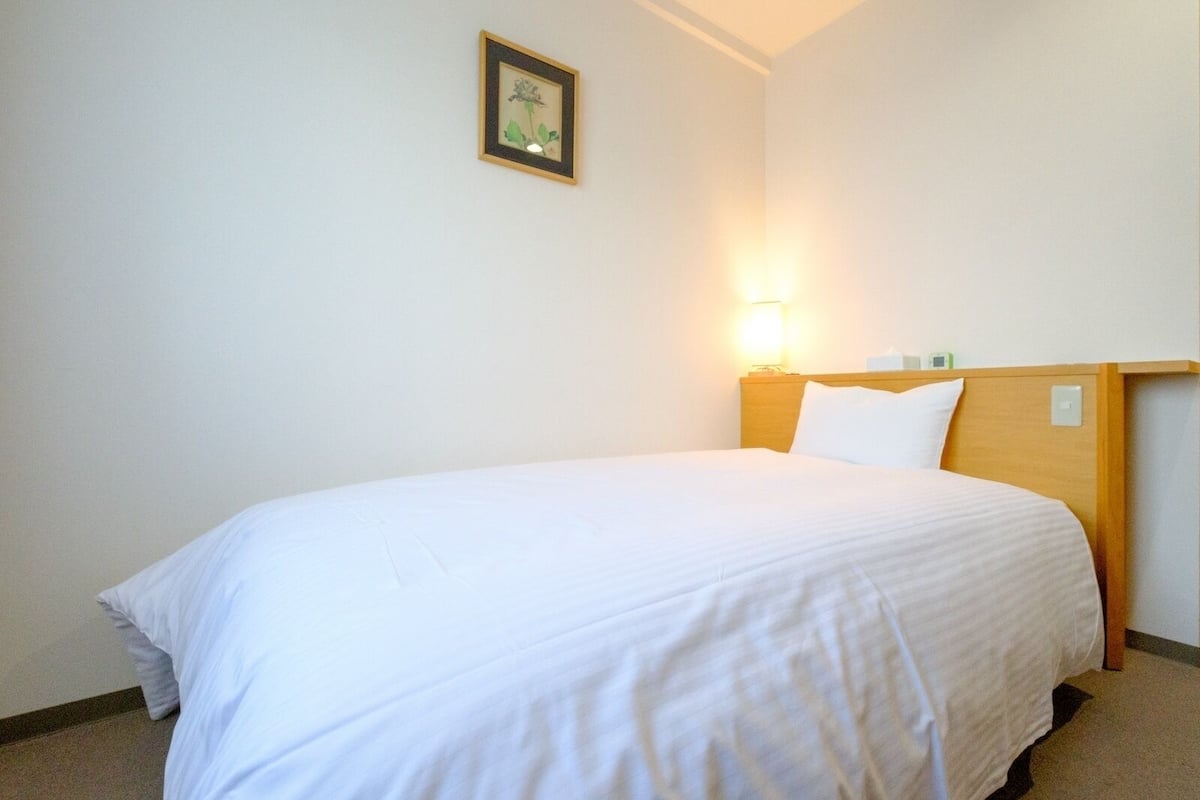 Een enkel bed met een houten hoofdbord, een nachtkastje met een lamp en een lijst met een afbeelding aan de muur.