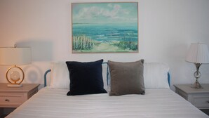 A hotel room with a bed, two pillows, a nightstand with a lamp, and a painting of a beach scene on the wall. - Relaxing 4-bedroom Pool Oasis Near Daytona (Port Orange)