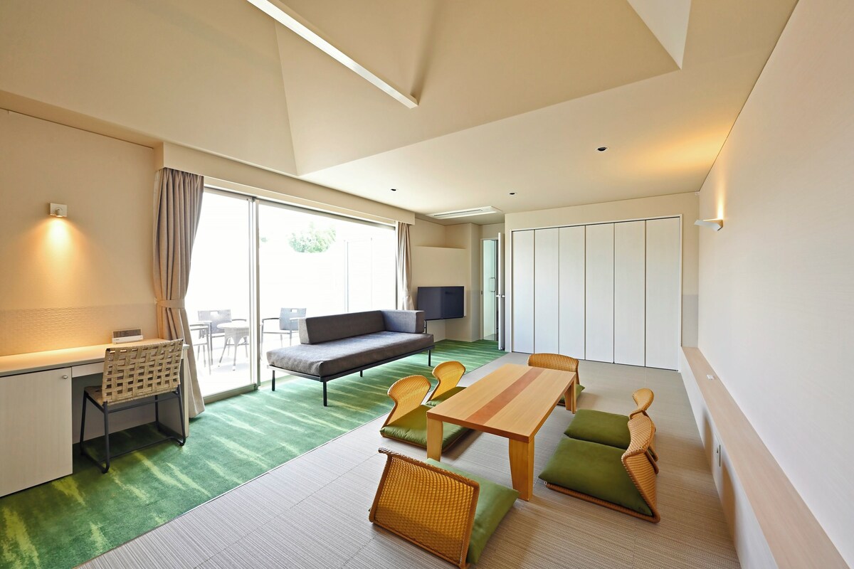 A modern hotel room with a sofa, dining table, chairs, a desk, and a TV.