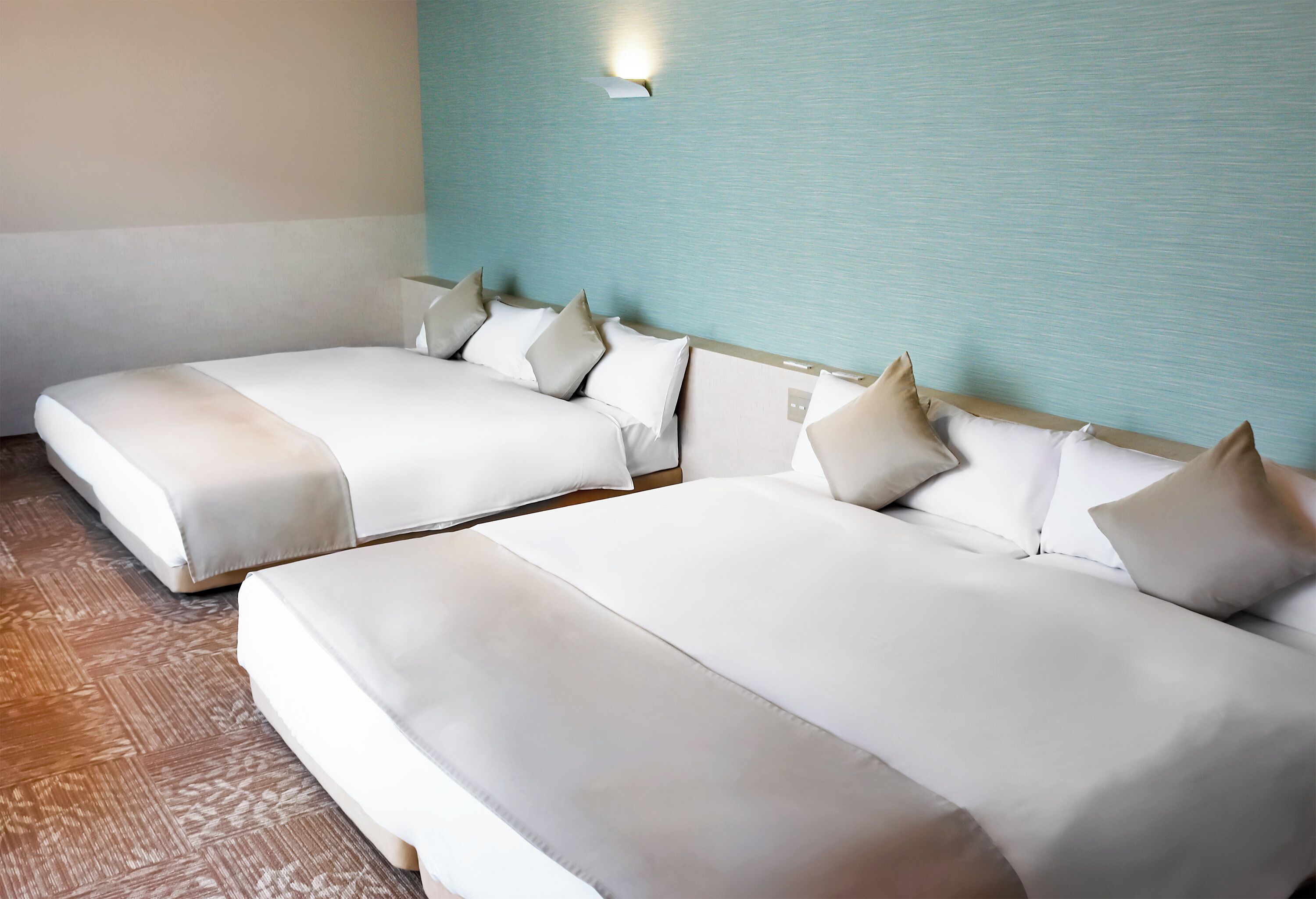 Two adjacent beds with white linens and pillows, a wooden floor, and a light blue wall with a wall-mounted light fixture.