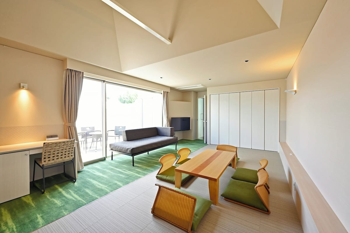 A modern hotel room with a sofa, dining table, chairs, a desk, and a TV.