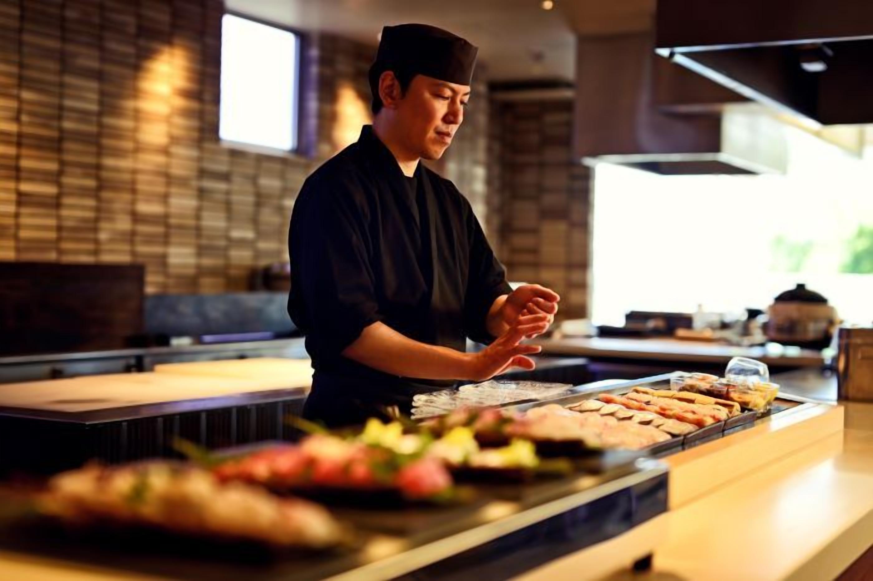 A sushi bar with a chef preparing food, a counter with various dishes, and a brick wall in the background.