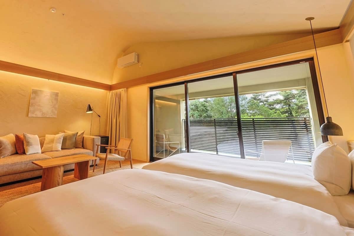 Une chambre d’hôtel avec un canapé, deux lits, une petite table, une lampe et une porte-fenêtre coulissante donnant sur un espace extérieur.