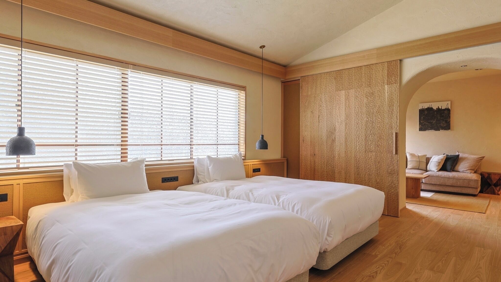 Camera da letto con due letti, pavimento in legno, una testiera di legno, una lampada a sospensione e un divano nell'angolo.