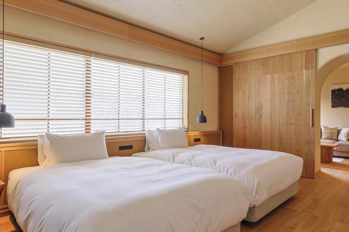 Une chambre avec deux lits, un plancher en bois, une tête de lit en bois, une lampe suspendue et un canapé placé dans un coin.