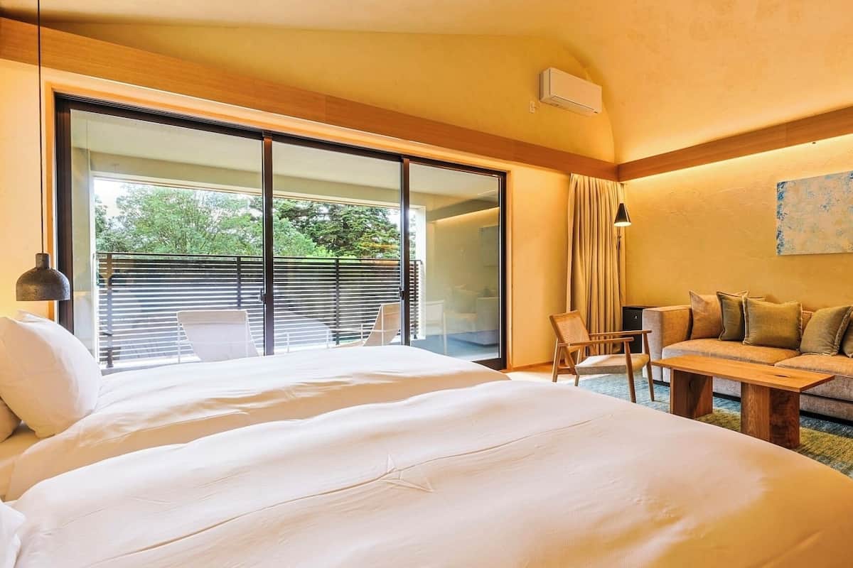 Une chambre d’hôtel avec un grand lit, un canapé, une table à manger, une chaise, une porte-fenêtre coulissante donnant sur un balcon et un tableau au mur.