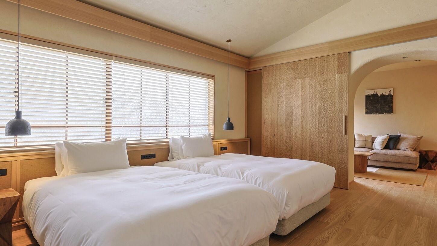 A hotel room with two beds, a wooden floor, a large window with blinds, a wall-mounted lamp, and a small sofa in the corner.