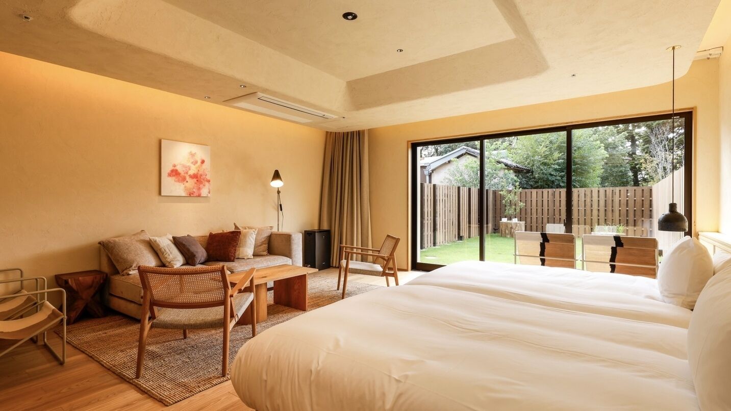 A modern hotel room with a large bed, a sofa, a dining table, and a view of the outdoor area.