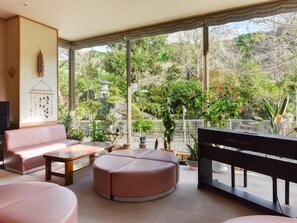 Un salon comprenant un canapé, un pouf rond, une table basse et un banc. De grandes fenêtres offrent une vue sur la verdure environnante.