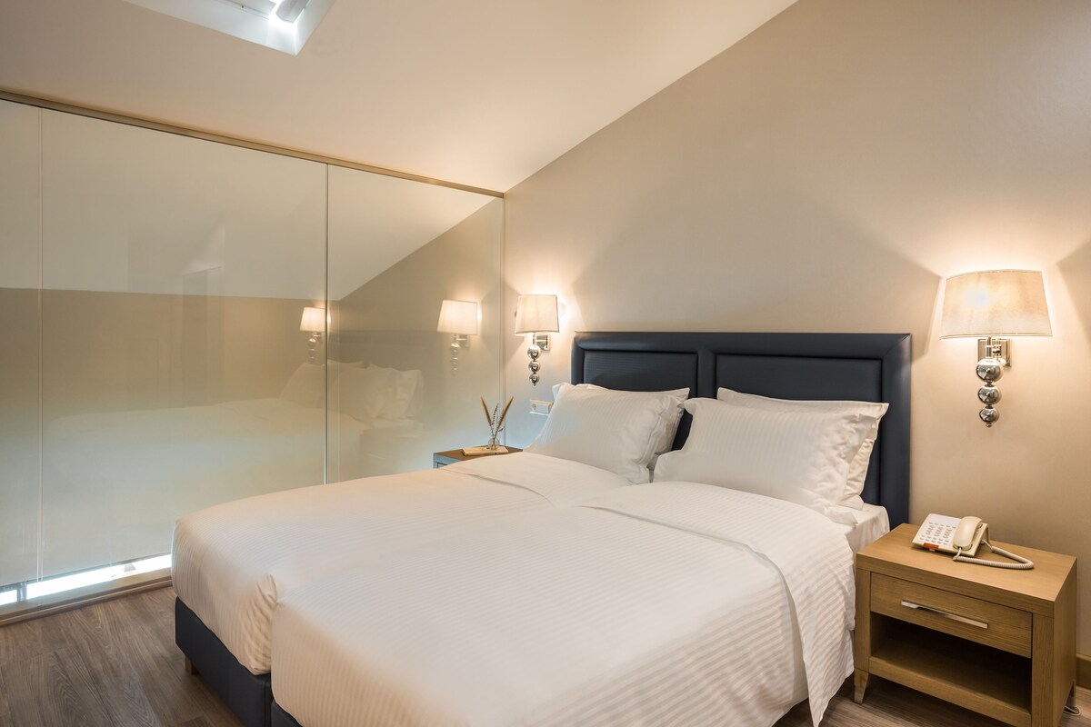 A modern hotel room with a large bed, bedside lamps, a nightstand, and a glass partition wall.