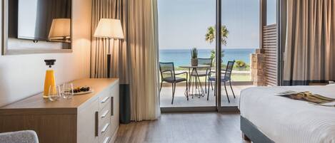 A hotel room with a bed, a desk with a lamp, a TV, a balcony with a table and chairs, and a view of the ocean.