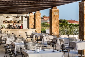 Ein Außenbereich mit Essbereich, Tischen, Stühlen, einer Bar mit Barkeeper und Blick auf ein Gebäude mit roten Dächern.