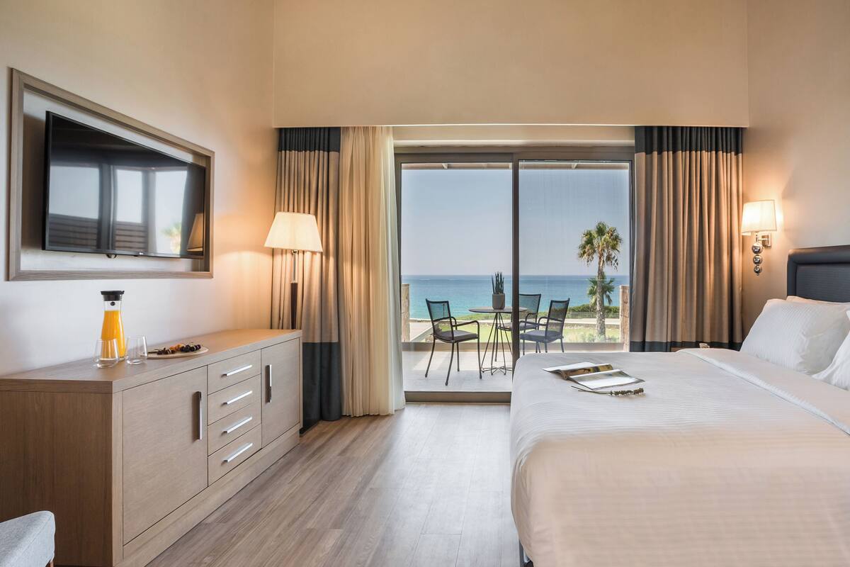 A hotel room with a bed, a TV, a desk with a lamp, a view of the ocean, and a balcony with chairs.