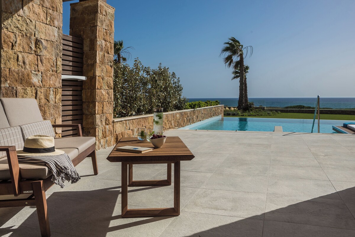 A patio with a pool, stone walls, a wooden table, and a lounge chair.