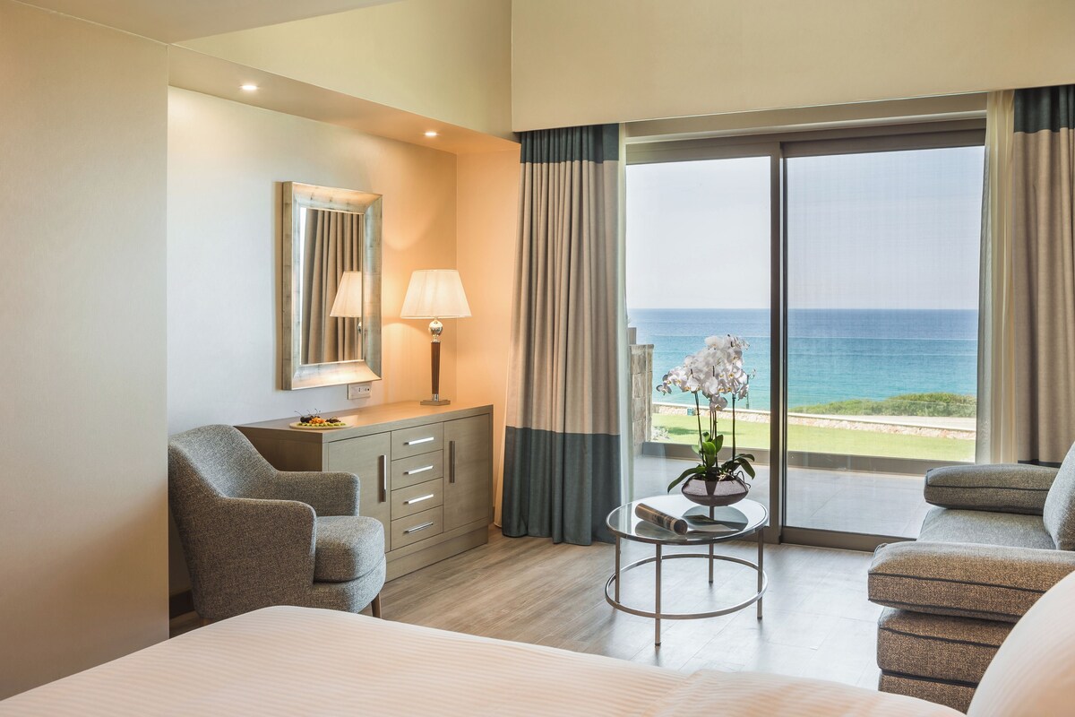 A hotel room with a bed, two armchairs, a small table with a lamp, a mirror, and a view of the ocean.