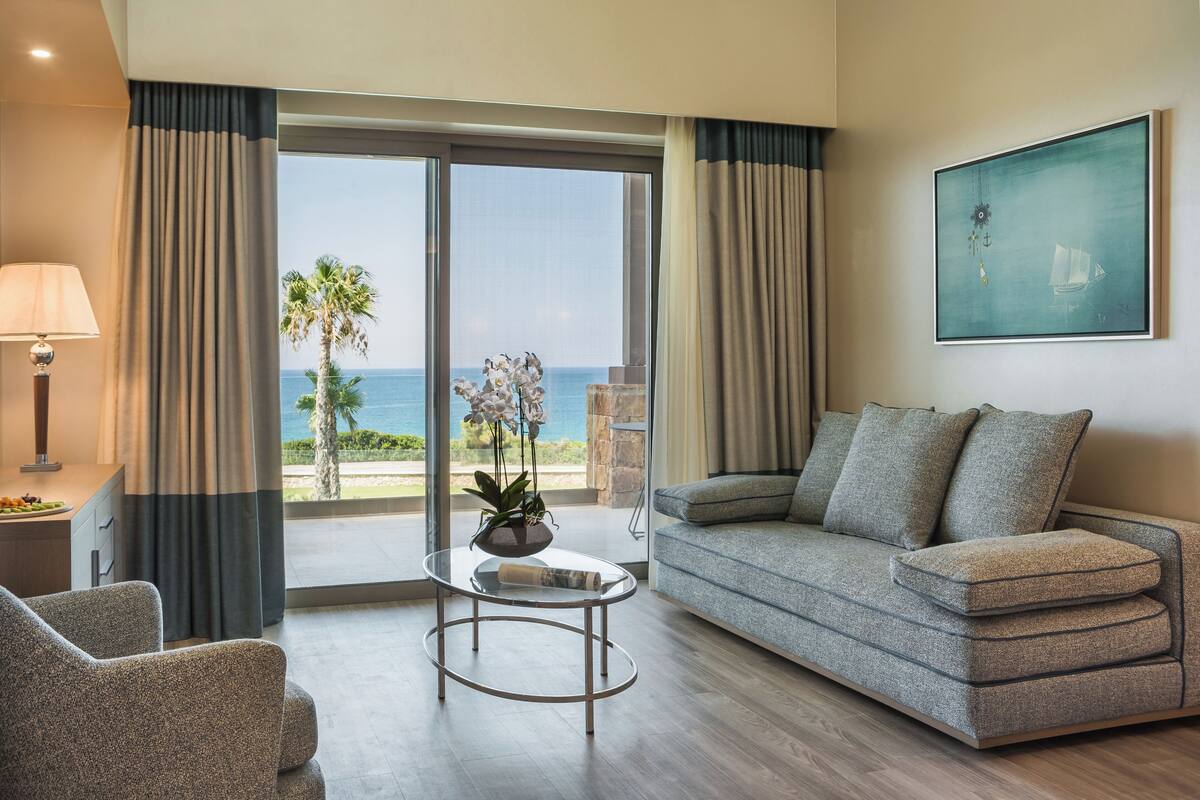 A modern living room with a sofa, armchair, and coffee table, a view of the ocean, and a painting on the wall.
