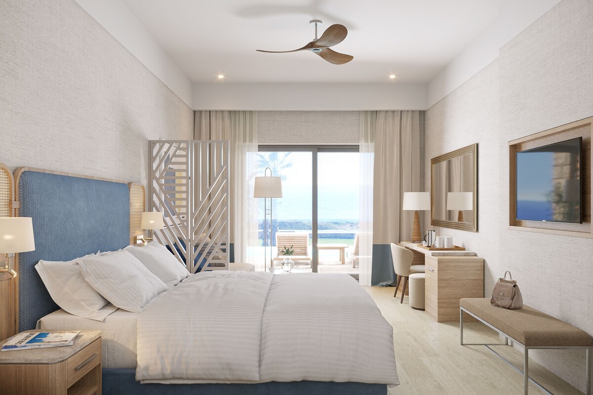 A modern hotel room with a large bed, a desk, a chair, a TV, and a balcony with a view.