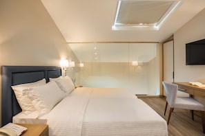 Una habitación de hotel con cama, mesita de noche, lámpara, escritorio, silla y televisor fijado a la pared.