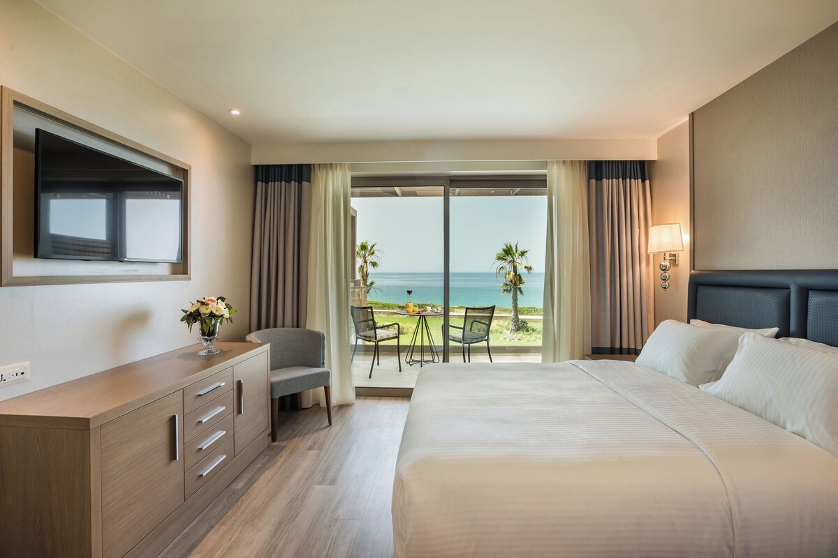A hotel room with a bed, a desk, a chair, a TV, and a balcony with a view of the ocean.