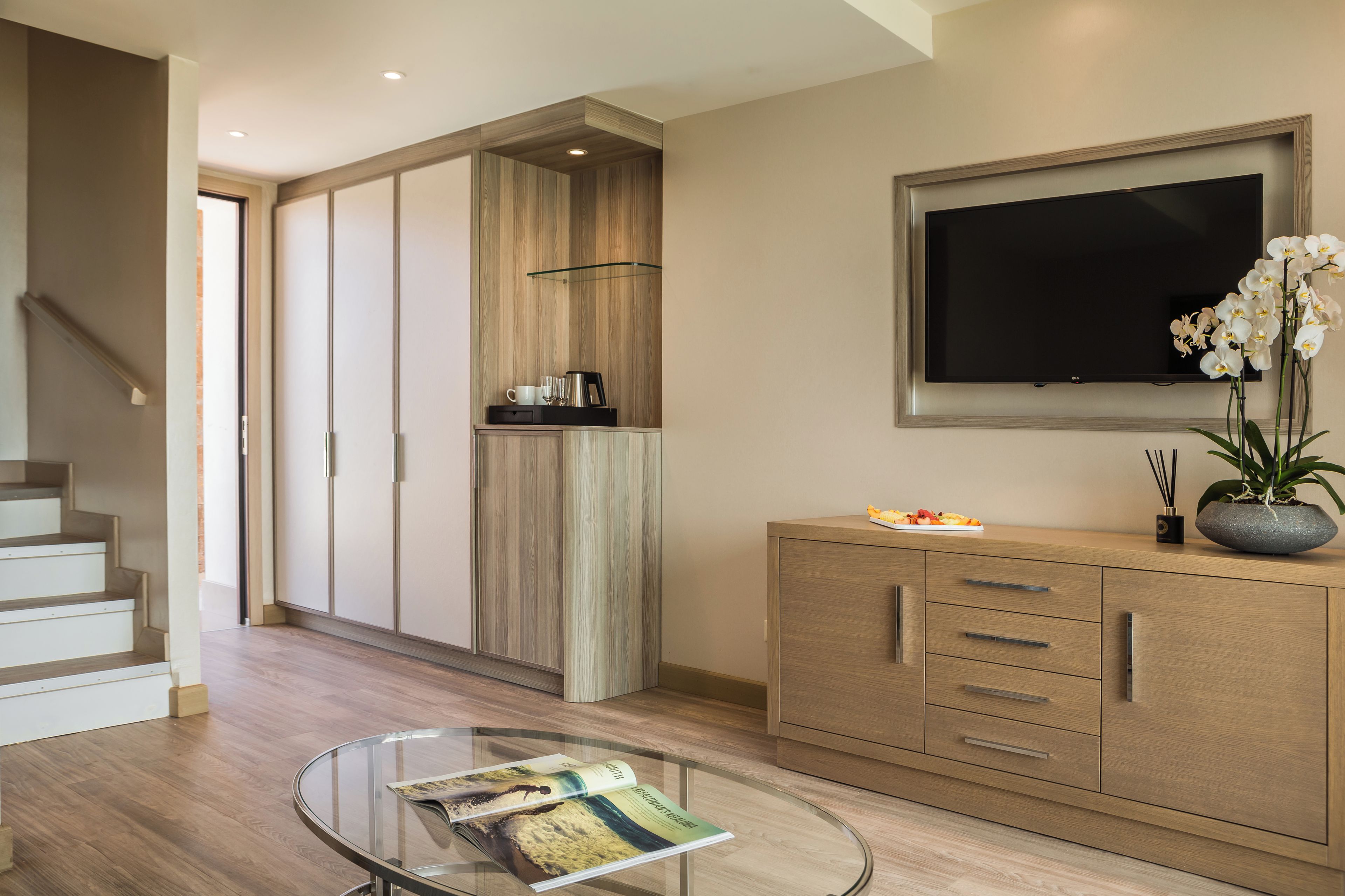 A modern living room with a glass coffee table, a flat-screen TV mounted on the wall, and a wooden cabinet with drawers.