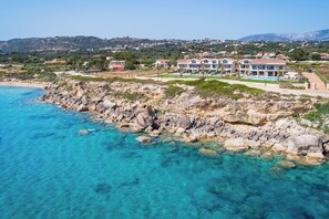 A coastal property with a rocky shoreline, clear blue water, and a view of a residential area in the background. - Electra Kefalonia Hotel and Spa (Kefalonia)