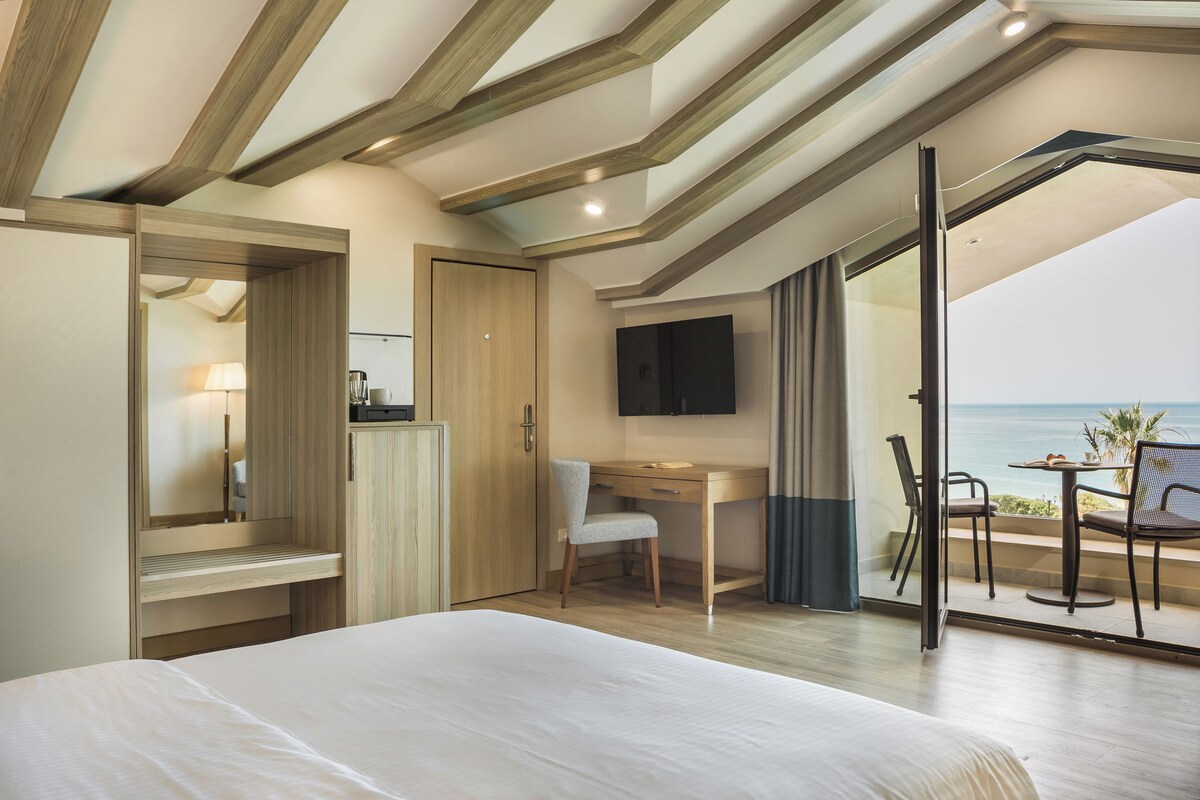 A bedroom with a bed, a desk, a chair, a TV, and a balcony with a view of the sea.