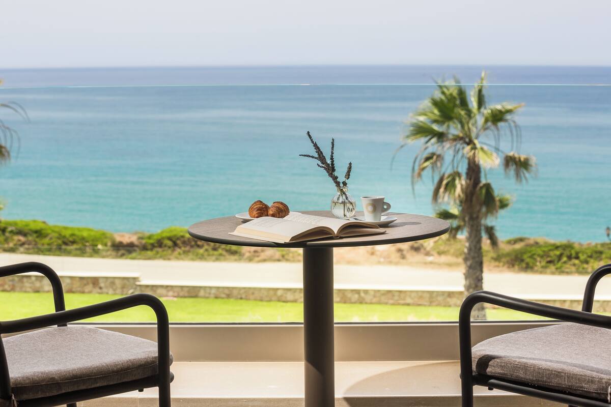 A table with a book, two croissants, and a vase with dried plants, overlooking a beach with palm trees and the ocean.