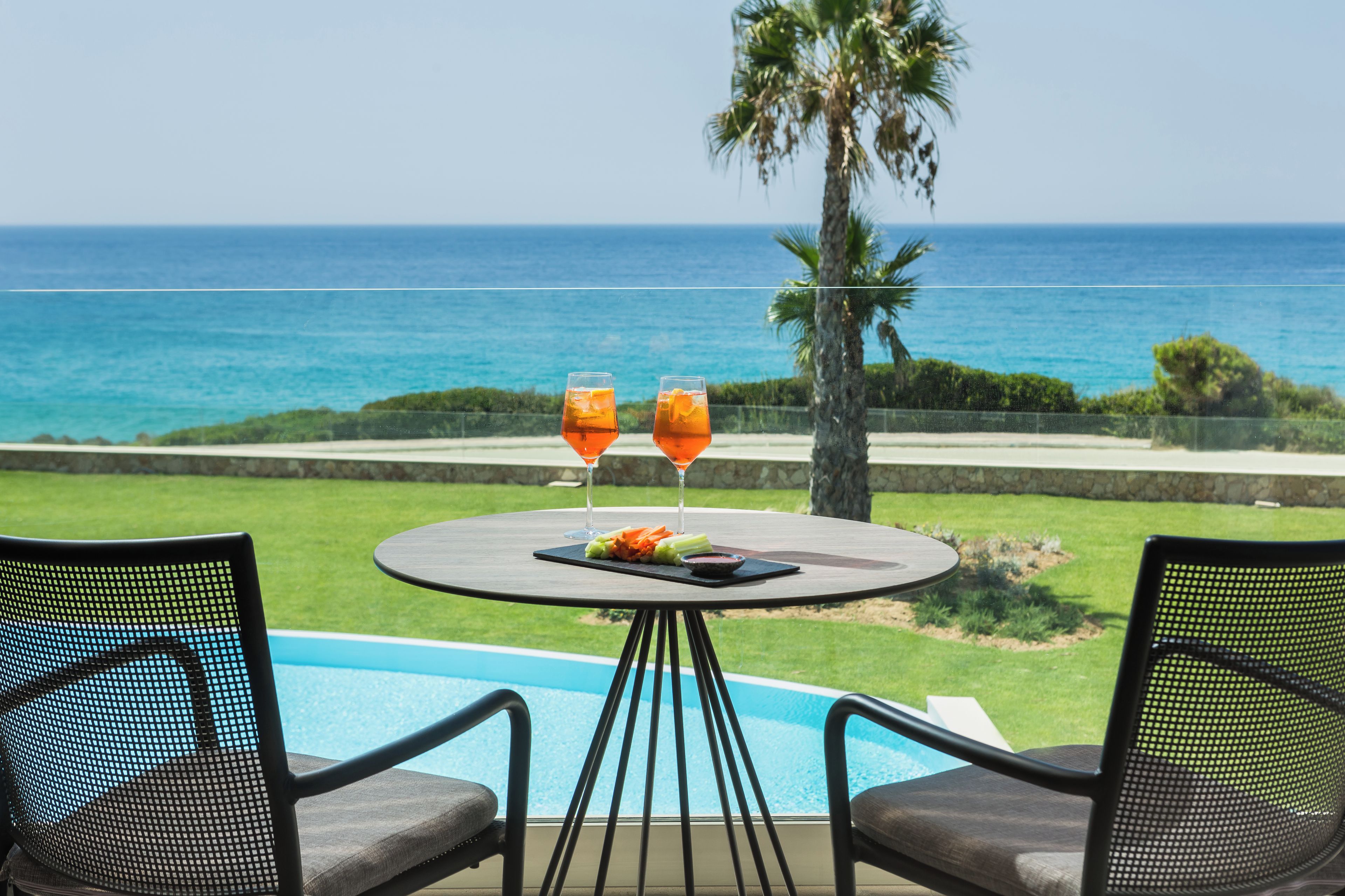 A poolside table with two glasses of orange beverage, a plate of food, and a view of the ocean.