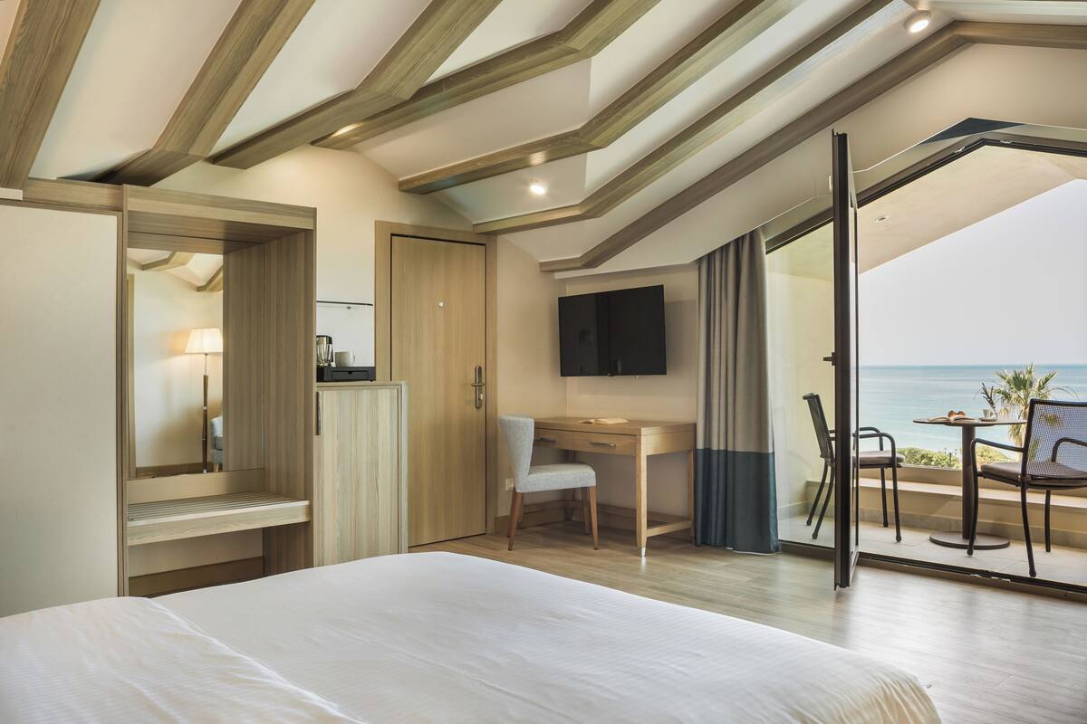 A bedroom with a bed, a desk, a chair, a TV, and a balcony with a view of the sea.