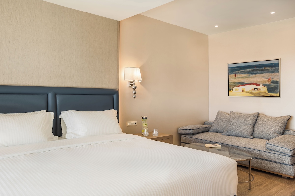 A hotel room with a bed, a sofa, a nightstand with a lamp, and a painting on the wall.
