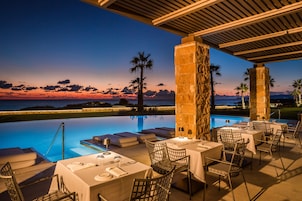 Un área de comedor al aire libre con mesas y sillas, una piscina y vista al atardecer sobre el océano.