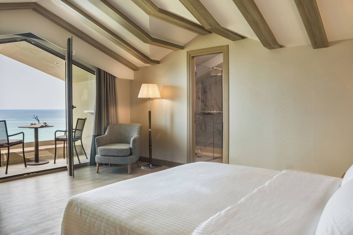 A hotel room with a bed, a chair, a table, a lamp, and a glass door leading to a balcony with a view of the sea.