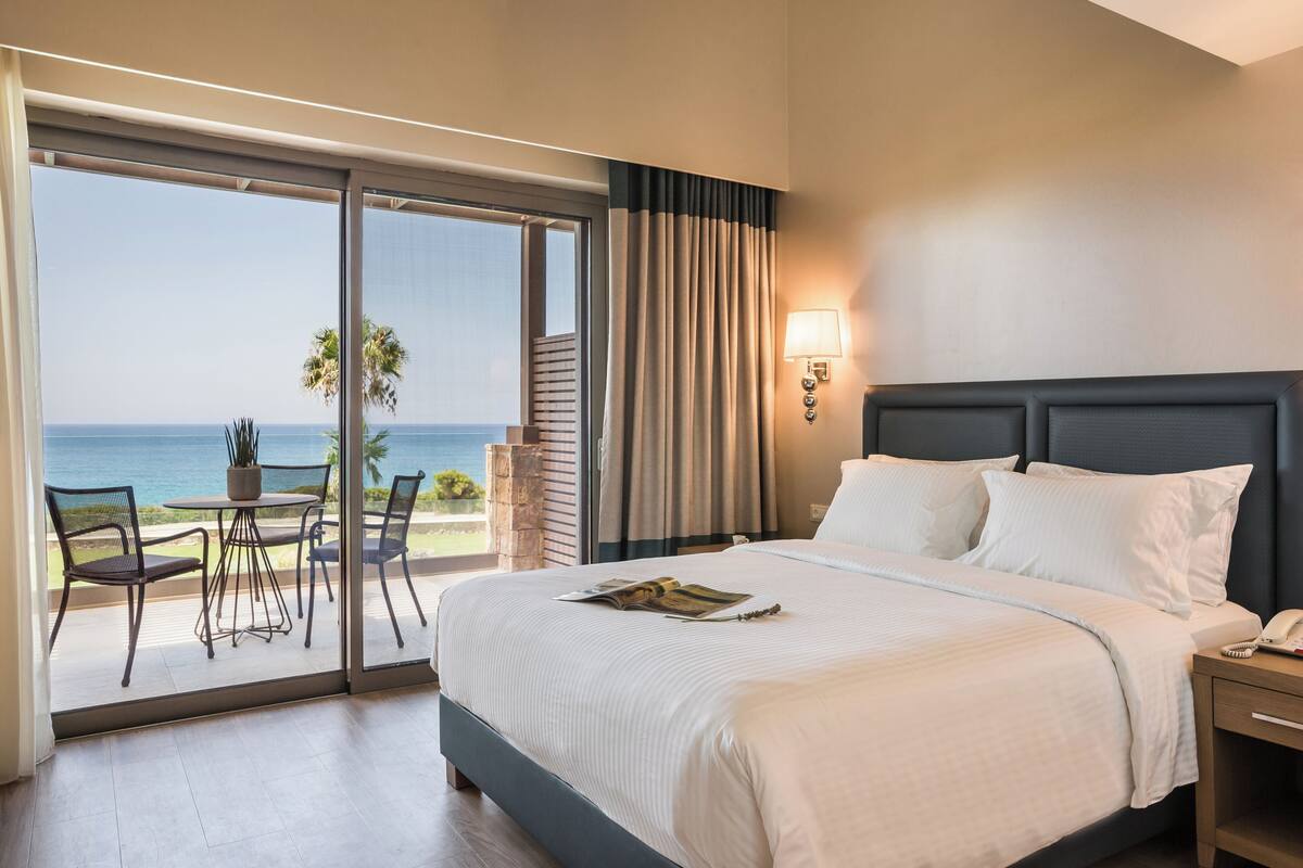 A hotel room with a bed, a bedside table, a lamp, a window with curtains, and a view of the ocean.