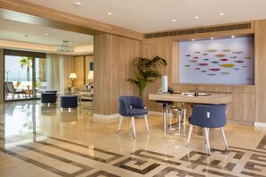 Un moderno vestíbulo de hotel con un mostrador de recepción, dos sillas azules, una planta grande en maceta y un cuadro de peces.