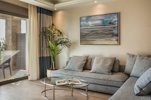 A modern living room with a sectional sofa, a glass coffee table with a magazine, a large abstract painting, and a potted plant. - Electra Kefalonia Hotel and Spa (Kefalonia)