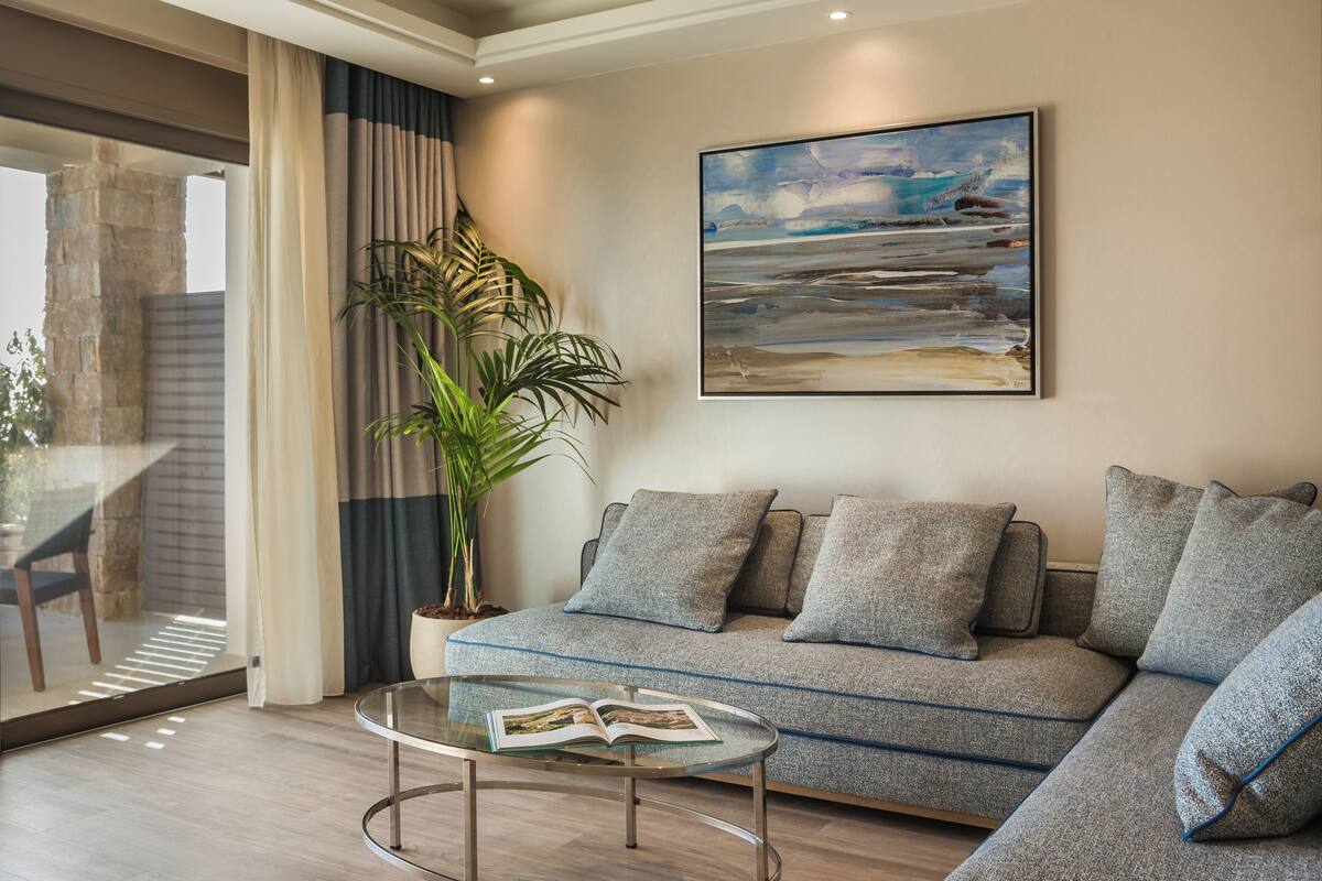 A modern living room with a sectional sofa, a glass coffee table with a magazine, a large abstract painting, and a potted plant.