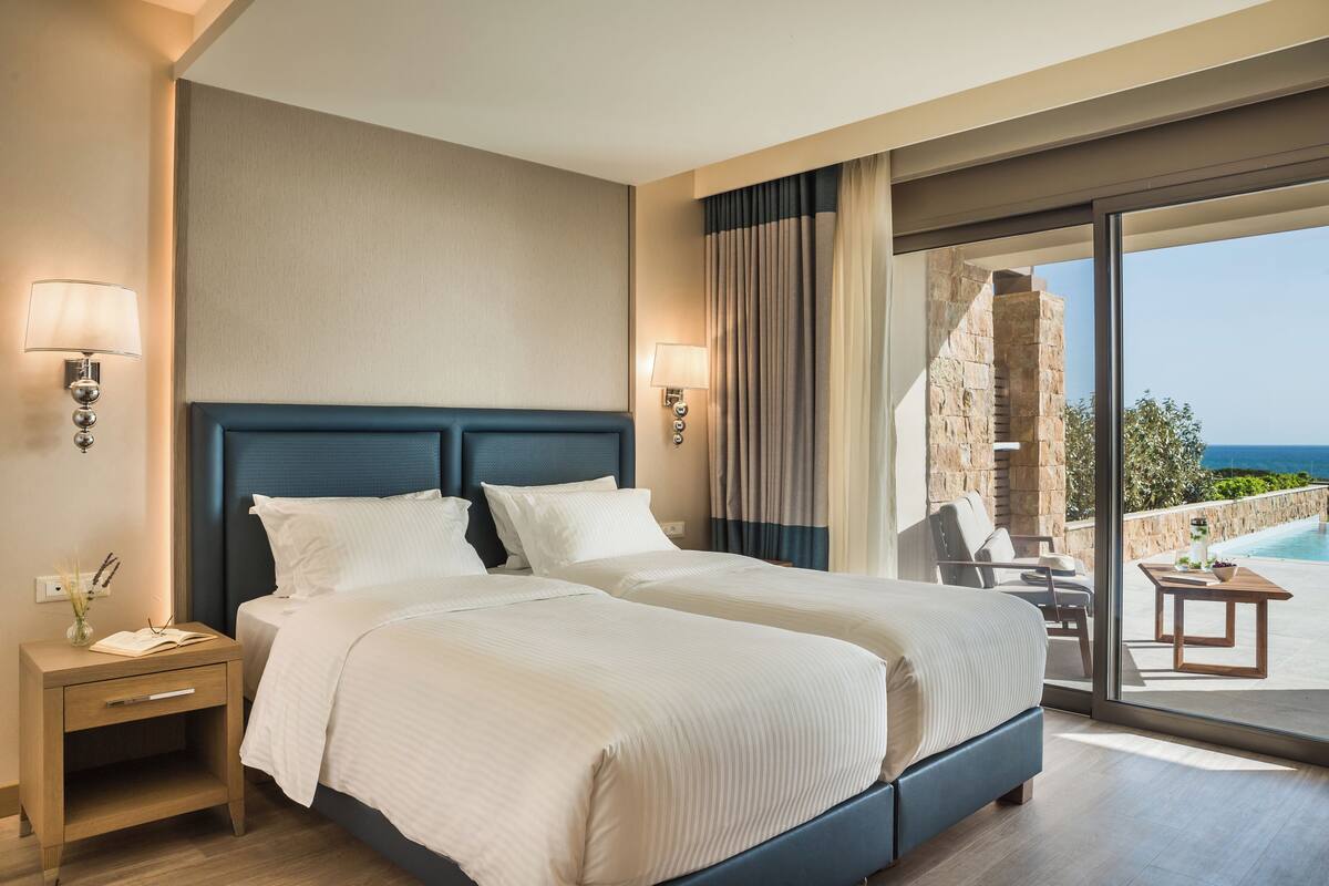 A hotel room with a large bed, two bedside tables, a nightstand, a view of the ocean, and a pool.