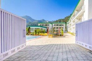A hotel entrance with a paved pathway leading to a pool area, surrounded by greenery and mountains in the background. - Alaya Stays By Elivaas Kingdom Heartz (Nainital)