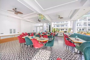 Un comedor moderno con suelo de diseño geométrico, varias mesas con sillas, ventiladores de techo y grandes ventanales que ofrecen vistas a un edificio y follaje.