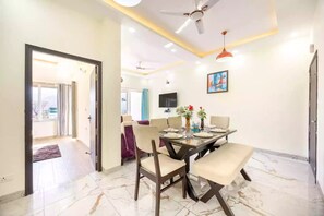 A dining area with a table set for four, a ceiling fan, and a TV mounted on the wall. - Alaya Stays By Elivaas Kingdom Heartz (Nainital)