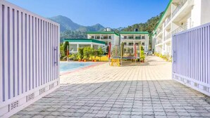 A hotel entrance with a paved pathway leading to a pool area, surrounded by greenery and mountains in the background. - Alaya Stays By Elivaas Kingdom Heartz (Nainital)