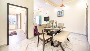 A dining area with a table set for four, a ceiling fan, and a TV mounted on the wall. - Alaya Stays By Elivaas Kingdom Heartz (Nainital)