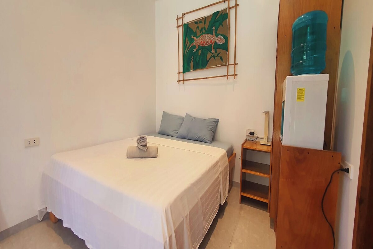 A small, neatly arranged bedroom with a bed, a nightstand, a water dispenser, and a wall-mounted artwork.