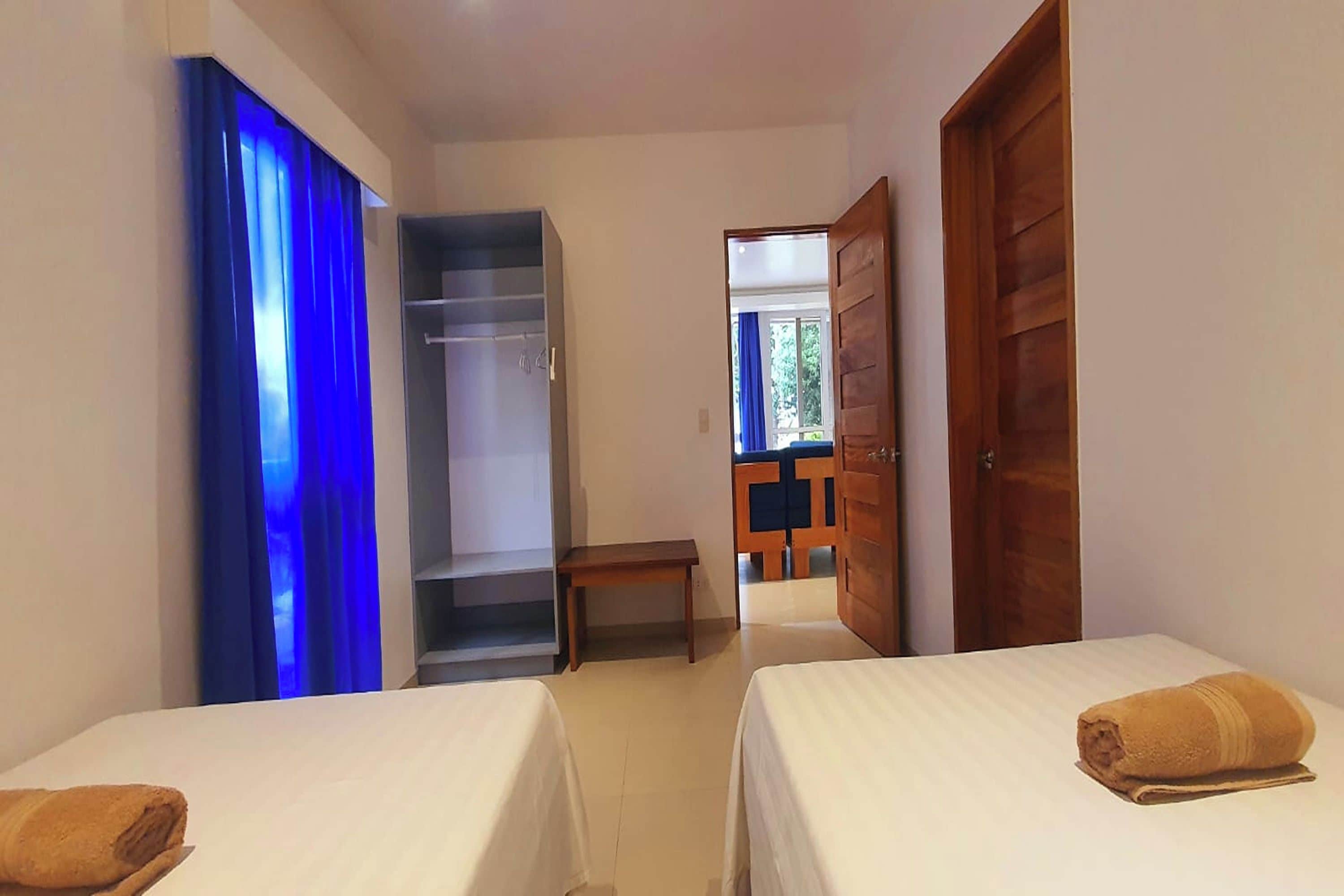 A hotel room with two beds, a wardrobe, a bench, and a view of the outside.