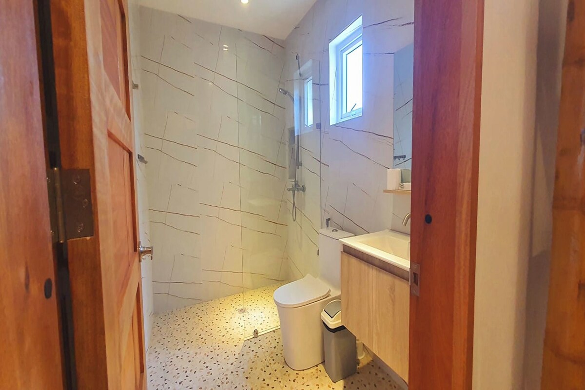 Ein Badezimmer mit einer Duschwand aus Marmor, einer Toilette, einem Mülleimer und einem Holzlagerflansch mit Spiegel.