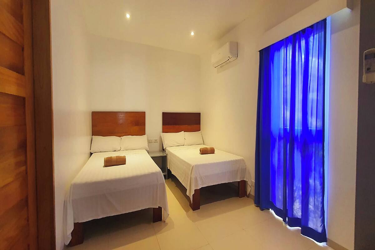 Une chambre d’hôtel avec deux lits simples, une tête de lit en bois, du linge de lit blanc et un rideau bleu.